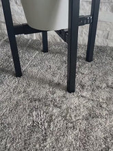 Load and play video in Gallery viewer, A beautifully composed lifestyle photograph in a sunlit modern living room, featuring the 2-tier black metal plant stand styled with a thriving snake plant in a white pot, placed next to a textured accent chair
