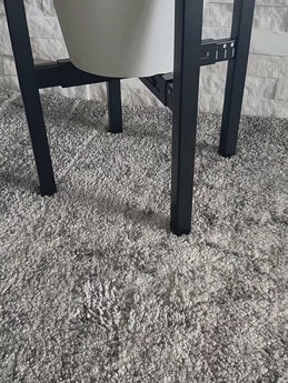 A beautifully composed lifestyle photograph in a sunlit modern living room, featuring the 2-tier black metal plant stand styled with a thriving snake plant in a white pot, placed next to a textured accent chair